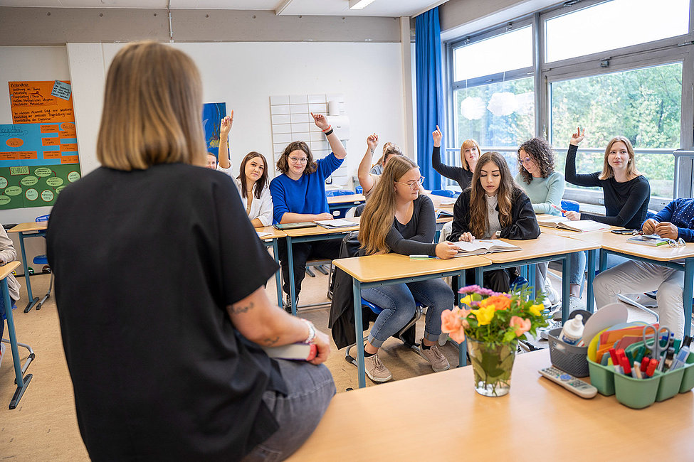 Lehrkraft von hinten im Klassenraum mehrere Studierende heben die Hand